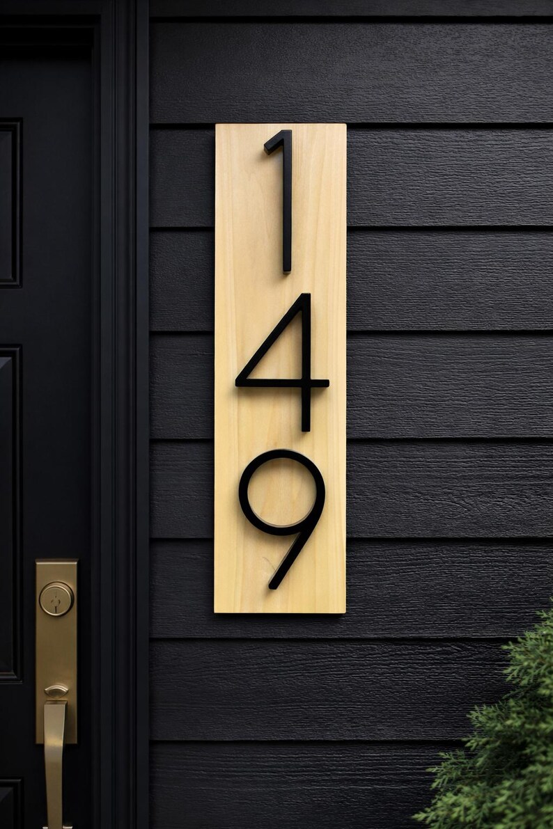 A Vertical modern house number sign featuring the modern black metal numbers "149", Mounted onto a light tone wooden rectangular 18 in x 5.5 in x 1/2 in thick plaque. The sign is set against a dark gray exterior wall. Potted plants below on the patio to the right of the address sign and a black front door with brass handle to the left.