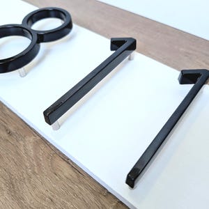 A close up side view of a Horizontal modern house number sign featuring the modern black metal numbers "811", shown floating mounted (intentionlly lifted to create space between numbers and plaque) onto a wooden white rectangular 18 in x 5.5 in x 1/2 in thick plaque.