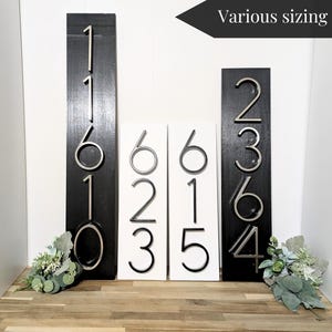 4 Vertical modern house number sign featuring the black and silver modern metal numbers, Mounted onto a wooden white and black rectangular plaques.  2 Plaques are 18 in x 5.5 in x 1/2 in thick 2 other plaques displayed are 24 in x 5.5 in x 1/2 in thick. All next to one another against a white wall.