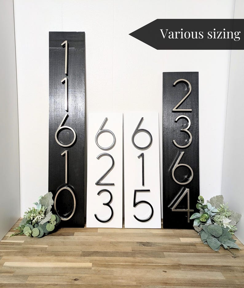 4 Vertical modern house number sign featuring the black and silver modern metal numbers, Mounted onto a wooden white and black rectangular plaques.  2 Plaques are 18 in x 5.5 in x 1/2 in thick 2 other plaques displayed are 24 in x 5.5 in x 1/2 in thick. All next to one another against a white wall.