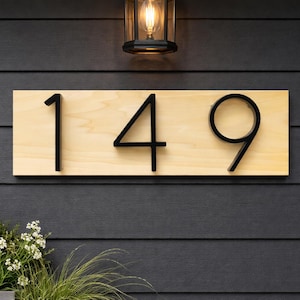 A Horizontal modern house number sign featuring the modern black metal numbers "149", Mounted onto a light wooden rectangular 18 in x 5.5 in x 1/2 in thick plaque. The sign is set against a black exterior wall. A small light is above the Horizontal adress sign while the top of green plants are peaking into the image from below the address sign.