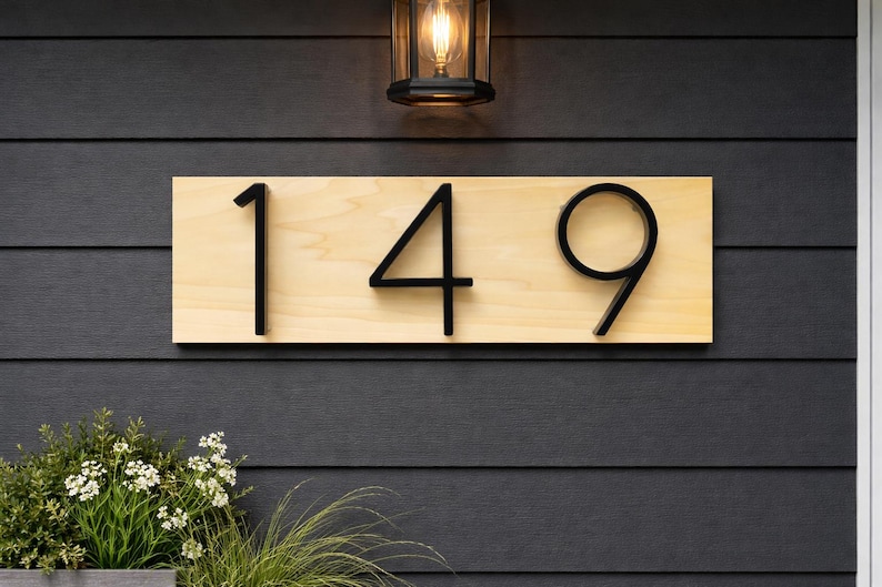 A Horizontal modern house number sign featuring the modern black metal numbers "149", Mounted onto a light wooden rectangular 18 in x 5.5 in x 1/2 in thick plaque. The sign is set against a black exterior wall. A small light is above the Horizontal adress sign while the top of green plants are peaking into the image from below the address sign.