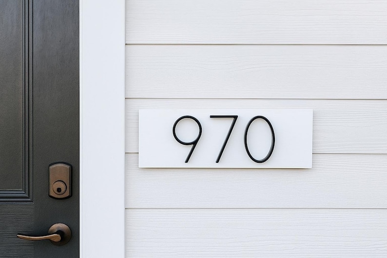 A Horizontal modern house number sign featuring the modern black metal numbers "970", Mounted onto a wooden white rectangular 18 in x 5.5 in x 1/2 in thick plaque. The sign is set against a white exterior wall. The sign is next to a dark gray door with a bronze handle and lock.