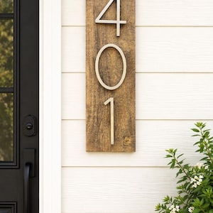 A Vertical modern house number sign featuring the modern silver metal numbers "401", Mounted onto a wooden warm honey stained rectangular 18 in x 5.5 in x 1/2 in thick plaque. The sign is set against a cream exterior wall. Potted plants below on the patio to the right of the address sign and  a black front door to the left.