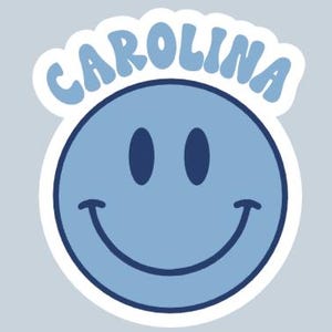 Peut inclure: Un autocollant de visage souriant bleu clair avec le mot "CAROLINA" dans une police bleu clair assortie au-dessus du visage. L'autocollant a une bordure blanche et est pos&eacute; sur un fond gris clair.