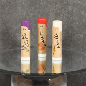 May include: Three lip balm tubes with different scents: Lavender Lemongrass, Strawberry, and Vanilla. The tubes are on a glass pedestal with a clear glass sphere filled with small black stones.