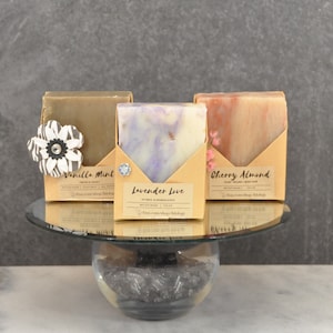 May include: Three bars of handmade soap on a mirrored cake stand. The soaps are wrapped in brown paper with labels that read "Vanilla Mint", "Lavender Love", and "Cherry Almond".