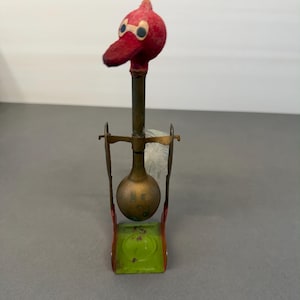 May include: A vintage toy featuring a red bird head with blue eyes atop a brass stem and a brass bulb. The bird is mounted on a green and red metal base with a wire frame. The toy has a whimsical, antique aesthetic.