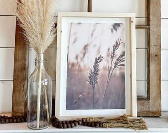 May include: A framed print of a sepia toned photograph of tall grasses in a field. The print is sitting on a white mantel with a vase of dried pampas grass and a brown beaded garland.