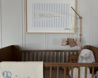 May include: A nursery scene featuring a wooden crib with a brown and white checkered mattress. A white blanket embroidered with the name "Briggs" in blue is draped over the side. A framed print with the text "Briggs Lane" hangs above the crib.