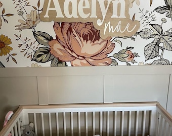 May include: A white crib with a floral patterned sheet and a white blanket. A stuffed animal sits on the sheet. A wooden sign with the name "Adelyn Mae" is hanging on the wall above the crib. The wall is covered in floral wallpaper.