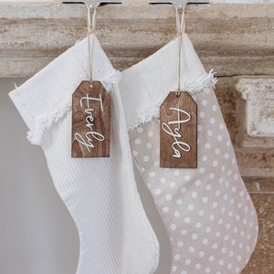 May include: Two Christmas stockings hang from silver clips on a mantel. One stocking is white with vertical stripes, the other is beige with white polka dots. Wooden tags with the names "Everly" and "Ayla" are attached.