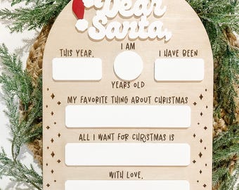 May include: Wooden Christmas sign with the words "Dear Santa" and space to write. The sign has a Santa hat and is surrounded by greenery. It includes fill-in-the-blank sections for personalizing a letter to Santa.