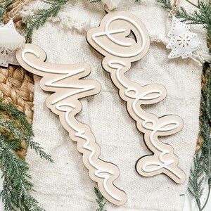 Puede incluir: Adornos de madera con las palabras "Mama" y "Dream" en fuente cursiva, delineadas en blanco. Los adornos son de color marrón claro y tienen una cuerda para colgar. El fondo incluye vegetación y una tela blanca.