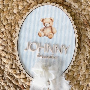 Puede incluir: Un letrero de madera con borde festoneado, con un oso de peluche de acuarela sobre un fondo a rayas azules y blancas. El nombre "JOHNNY" se muestra en letras de madera, con un nombre más pequeño debajo. Se adjunta un lazo de cinta blanca en la parte inferior.