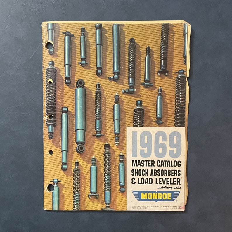 Pu&ograve; includere: Un catalogo vintage Monroe Master del 1969 per ammortizzatori e stabilizzatori di livello di carico. La copertina del catalogo presenta una variet&agrave; di ammortizzatori di diverse dimensioni e stili.