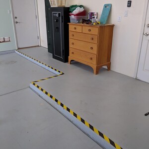 May include: A black safe with a yellow and black caution tape border on the floor. A wooden dresser with six drawers is to the right of the safe. The dresser is against a white wall.