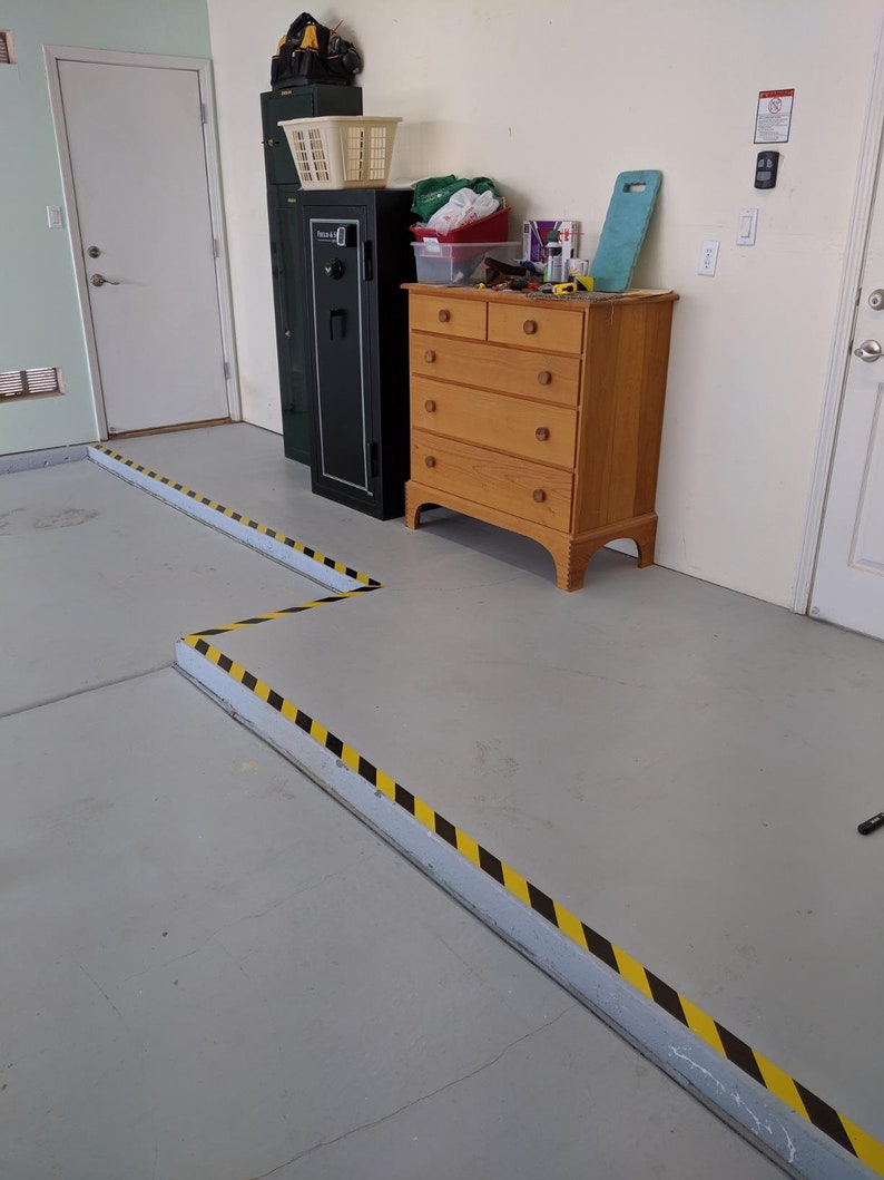 May include: A black safe with a yellow and black caution tape border on the floor. A wooden dresser with six drawers is to the right of the safe. The dresser is against a white wall.