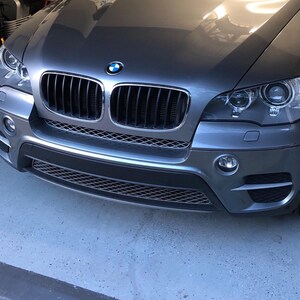 May include: A gray BMW X5 SUV with a black grille and headlights. The car is parked in a garage with yellow and black caution tape on the floor.