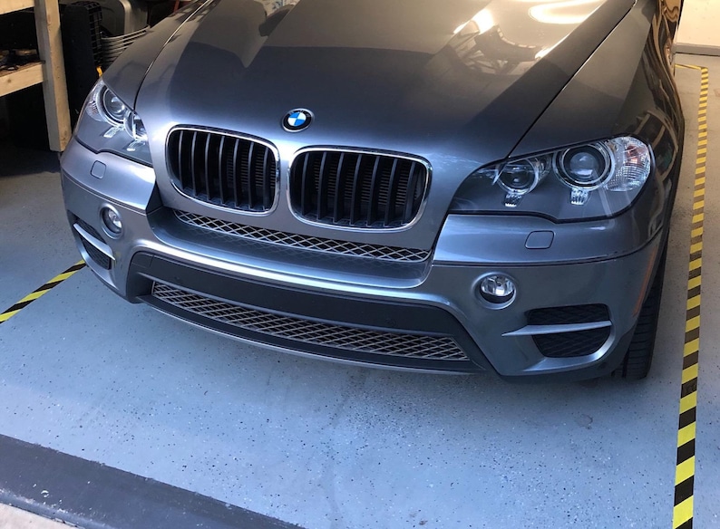 May include: A gray BMW X5 SUV with a black grille and headlights. The car is parked in a garage with yellow and black caution tape on the floor.