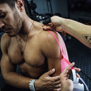May include: A person is applying pink kinesiology tape to a man's shoulder. The tape is being applied to the shoulder and upper arm. The person applying the tape has a tattoo of a lion's head in a triangle on their arm.