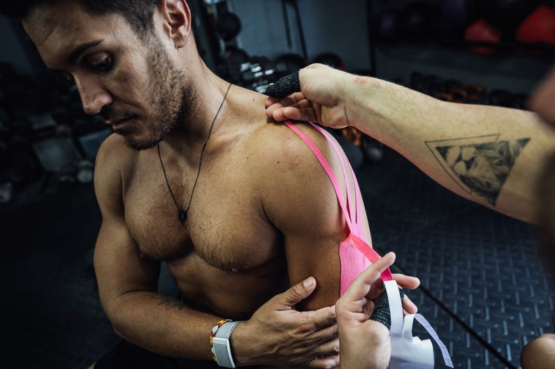 May include: A person is applying pink kinesiology tape to a man's shoulder. The tape is being applied to the shoulder and upper arm. The person applying the tape has a tattoo of a lion's head in a triangle on their arm.