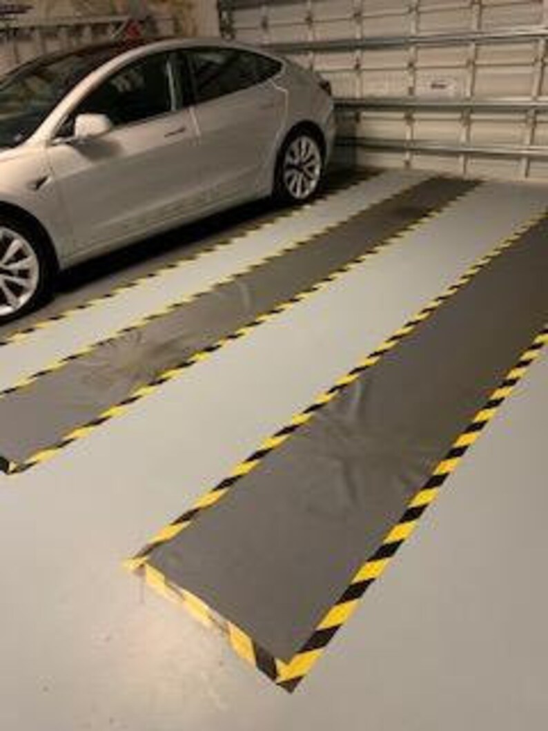 May include: Two gray garage floor mats with black and yellow caution tape markings. The mats are in a garage with a silver Tesla car parked in the background.