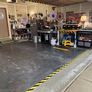 May include: A garage with a black and yellow striped floor, a black metal shelving unit, a black grill, a black and yellow striped table, a yellow and black cart, and a black and silver cart with shelves. The garage has a door with a Route 66 sign and a sign that says "BBQ".