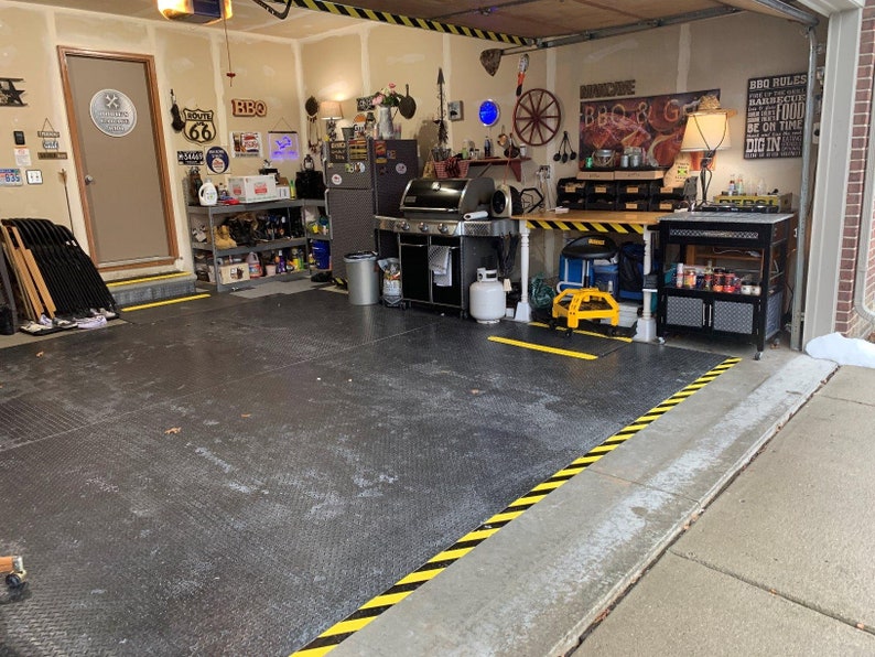 May include: A garage with a black and yellow striped floor, a black metal shelving unit, a black grill, a black and yellow striped table, a yellow and black cart, and a black and silver cart with shelves. The garage has a door with a Route 66 sign and a sign that says "BBQ".