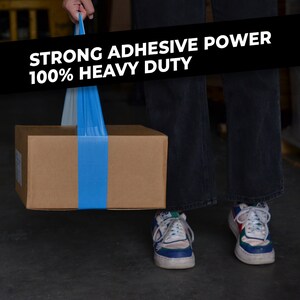 May include: A person is holding a brown cardboard box with a blue tape strap. The box is being lifted by the strap. The text on the strap reads "STRONG ADHESIVE POWER 100% HEAVY DUTY".