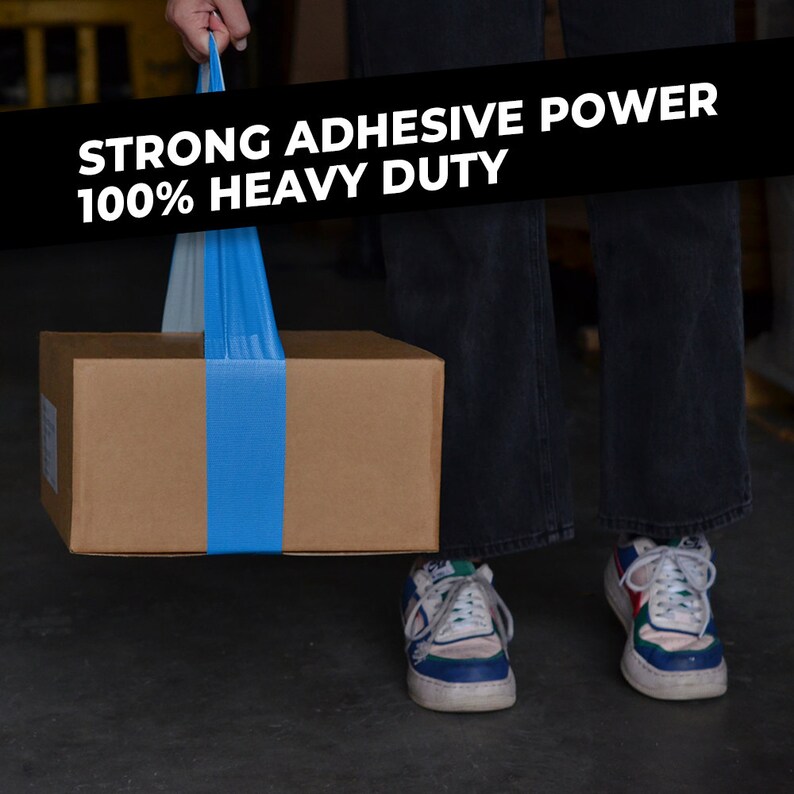 May include: A person is holding a brown cardboard box with a blue tape strap. The box is being lifted by the strap. The text on the strap reads "STRONG ADHESIVE POWER 100% HEAVY DUTY".