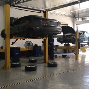 May include: A car repair shop with four cars on lifts. Two cars are lifted high enough to see the underside of the vehicles. Two cars are lifted lower, with the hoods open. There are four tires on the floor, and a yellow and black striped safety line on the floor.
