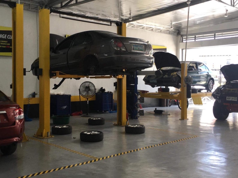 May include: A car repair shop with four cars on lifts. Two cars are lifted high enough to see the underside of the vehicles. Two cars are lifted lower, with the hoods open. There are four tires on the floor, and a yellow and black striped safety line on the floor.