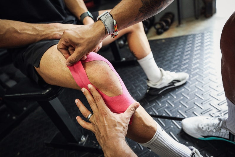 May include: A person's knee is being taped with pink athletic tape. The tape is being applied in a criss-cross pattern.
