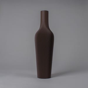 Könnte beinhalten: Eine hohe, braune Vase mit schmalem Hals und breitem Boden. Die Vase hat eine gerippte Textur mit vertikalen Linien, die von der Basis zum Hals verlaufen. Die Vase besteht aus einem festen Material und hat eine matte Oberfläche. Ein dekorativer Artikel.