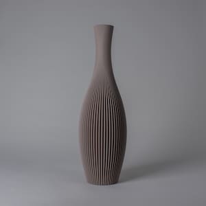 Può includere: Un vaso alto e slanciato di colore malva-grigio tenue. La superficie esterna presenta una consistenza a coste verticali che corre dal collo stretto fino alla base più ampia e arrotondata, creando un motivo uniforme e ripetitivo.
