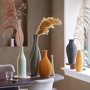 Op de afbeelding: Een verzameling decoratieve vazen in verschillende kleuren en texturen, waaronder een geribbelde oranje vaas, een getextureerde lichtgroene vaas en een zwarte vaas met zigzagpatroon. De vazen staan op een donkere houten tafel met een brandende kaars en decoratieve planten.