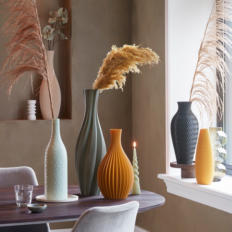 Op de afbeelding: Een verzameling decoratieve vazen in verschillende kleuren en texturen, waaronder een geribbelde oranje vaas, een getextureerde lichtgroene vaas en een zwarte vaas met zigzagpatroon. De vazen staan op een donkere houten tafel met een brandende kaars en decoratieve planten.