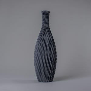 Op de afbeelding: Een hoge, donkergrijze decoratieve vaas met een textuur, geweven patroon. De vaas heeft een slanke hals en een bredere, afgeronde body. Het ontwerp heeft een herhalende, golfachtige textuur die het hele oppervlak bedekt.