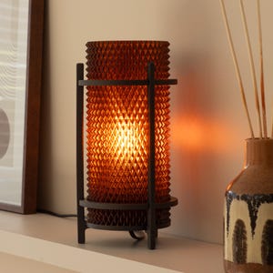 May include: A decorative table lamp with an amber-coloured, textured shade. The lamp is supported by a dark metal frame. The lamp is turned on, emitting a warm glow. The lamp is on a white surface.