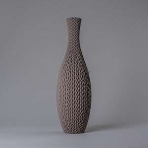 Könnte beinhalten: Eine hohe, taupefarbene Vase mit einem strukturierten, gewebten Muster. Die Vase hat einen schmalen Hals, der sich zu einem bauchigen Körper erweitert und sich dann zur Basis verjüngt. Die neutrale Farbe und die einzigartige Textur machen sie zu einem dekorativen Wohnakzent, ca. 30 cm hoch.