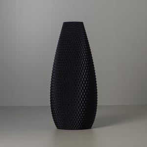 Könnte beinhalten: Eine hohe, schwarze Vase mit einer strukturierten, rautenförmigen Oberfläche. Die Vase hat einen breiten Boden, der sich nach oben verjüngt. Die Vase besteht aus einem mattschwarzen Material und eignet sich für die Inneneinrichtung.