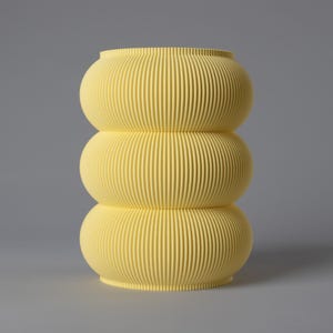 Op de afbeelding: Een gestapelde, gele decoratieve vaas met een geribbelde textuur. De vaas bestaat uit drie afgeronde secties, elk met verticale groeven, waardoor een uniek, modern ontwerp ontstaat. De vaas heeft een effen, lichtgele kleur en staat tegen een grijze achtergrond.