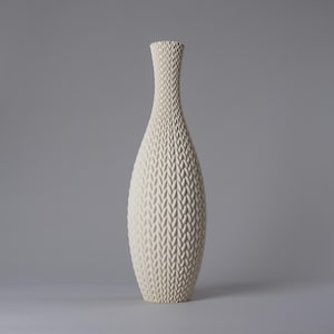 Può includere: Un vaso alto in ceramica bianco sporco con un motivo strutturato simile a un tessuto. Il vaso ha un collo sottile che si allarga in un corpo bulboso, poi si restringe verso la base. Il colore neutro e la trama unica lo rendono un pezzo decorativo.