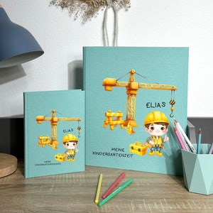 Könnte beinhalten: Zwei türkisfarbene Hardcover-Journale mit einem Bau-Thema. Das größere Journal hat den Text "Meine Kindergartenzeit" und "Elias" auf dem vorderen Umschlag. Das kleinere Journal hat den Text "Meine Kindergartenfreunde" und "Elias" auf dem vorderen Umschlag. Beide Journale zeigen eine Comic-Illustration eines Bauarbeiters mit gelbem Helm und einem gelben Baukran.