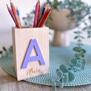 Peut inclure: Un porte-crayons en bois avec la lettre "A" et le nom "Alicia" gravés sur le devant. Le porte-crayons est rempli de crayons de couleur.