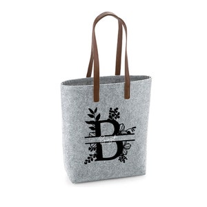 Könnte beinhalten: Graue Filztasche mit braunen Ledergriffen. Die Tasche hat ein schwarzes Blumenmuster mit dem Buchstaben "B" und dem Namen "Birgit" darauf gedruckt.