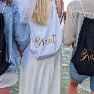 Może przedstawiać: Trzy osoby noszące czarne plecaki sznurkowe z złotymi napisami "Team Braut". Osoba w środku nosi białą sukienkę z welonem i złoty plecak sznurkowy z napisem "Braut".