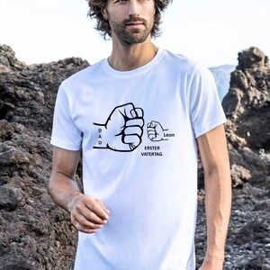 Pu&ograve; includere: Maglietta bianca con una grafica nera di due pugni che si scontrano. Il pugno pi&ugrave; grande dice "DAD" e il pugno pi&ugrave; piccolo dice "Leon". Il testo "ERSTER VATERTAG" &egrave; sotto i pugni.