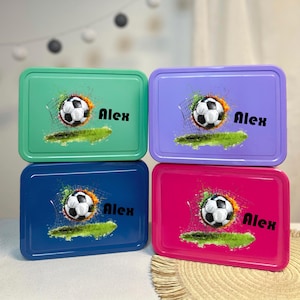 Peut inclure: Quatre boîtes à lunch rectangulaires en métal, vertes, violettes, bleues et roses. Chacune présente un motif de ballon de football avec un filet, de l'herbe et le nom "Alex" en noir. Les ballons sont noirs et blancs.
